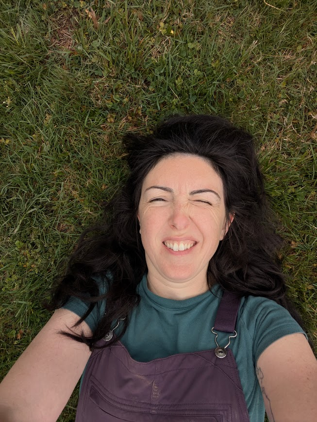 A photo of the author laying in the grass wearing purple overalls and squinting into the sunlight.